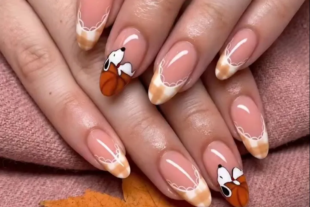 Almond nails with sheer pink base, orange gingham French tips edged in white lace, and a cartoon beagle on a pumpkin accent nail, hands resting on a pink knit sweater